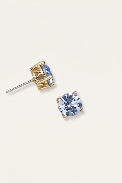 A pair of gold stud earrings with oval light blue gemstones, one shown from the front and the other from the side/back, on a light cream background.