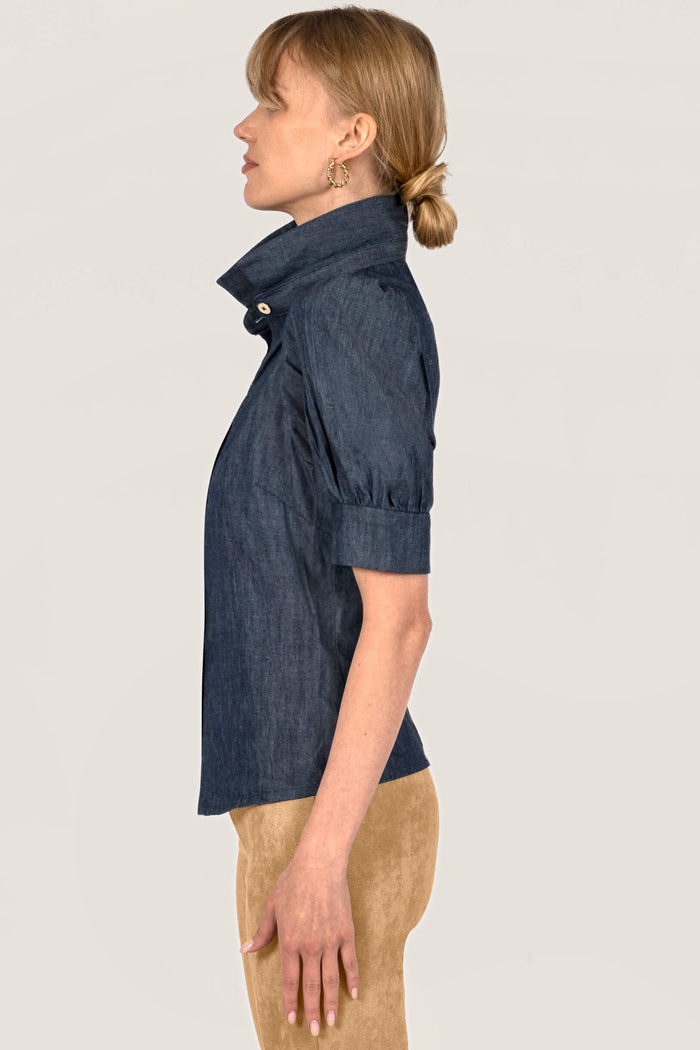 A woman shown in side profile wearing a dark indigo chambray short-sleeve blouse with a high collar and tan pants, set against a plain off-white background.
