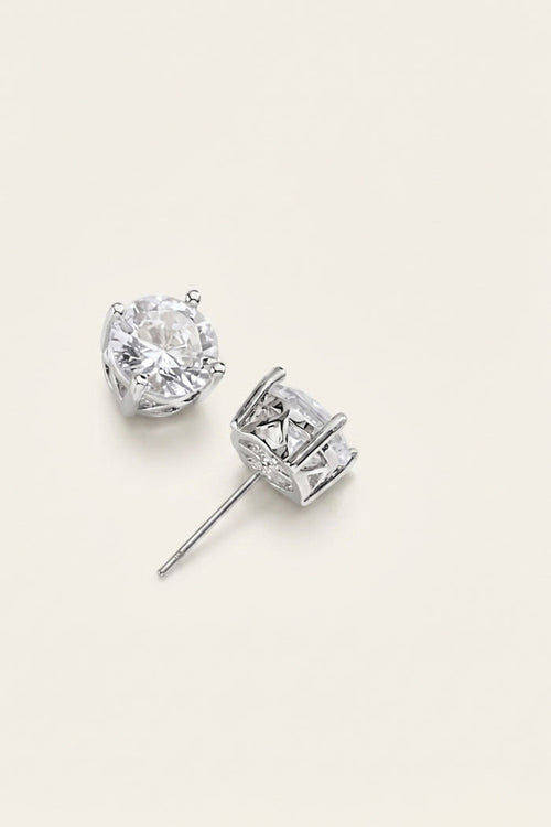 A pair of silver stud earrings with clear gemstones, one shown from the front and the other from the side, on a light beige background.