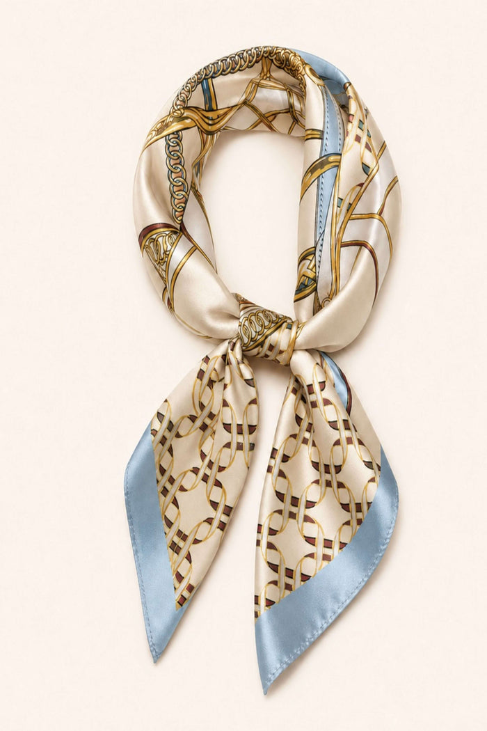 A silk scarf with a beige and gold geometric chain-link pattern and light blue edges, tied in a loop on a light cream background.