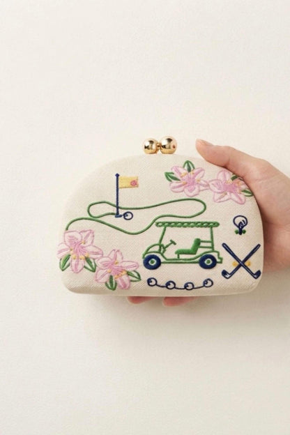 A hand holding a small cream-colored clutch purse decorated with embroidered golf-themed motifs, including a green golf cart, pink flowers, a flagstick, and crossed clubs, with a gold-tone clasp closure.