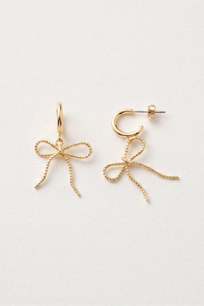 A pair of gold-toned earrings on a clean white background, featuring one dangling bow-shaped earring and one matching hoop earring with a small bow detail.
