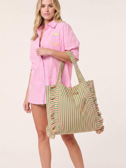 A woman models a pink button-up shirt dress and carries a large striped tote bag with green, yellow, and pink vertical stripes and ruffle trim, against a plain light beige background.